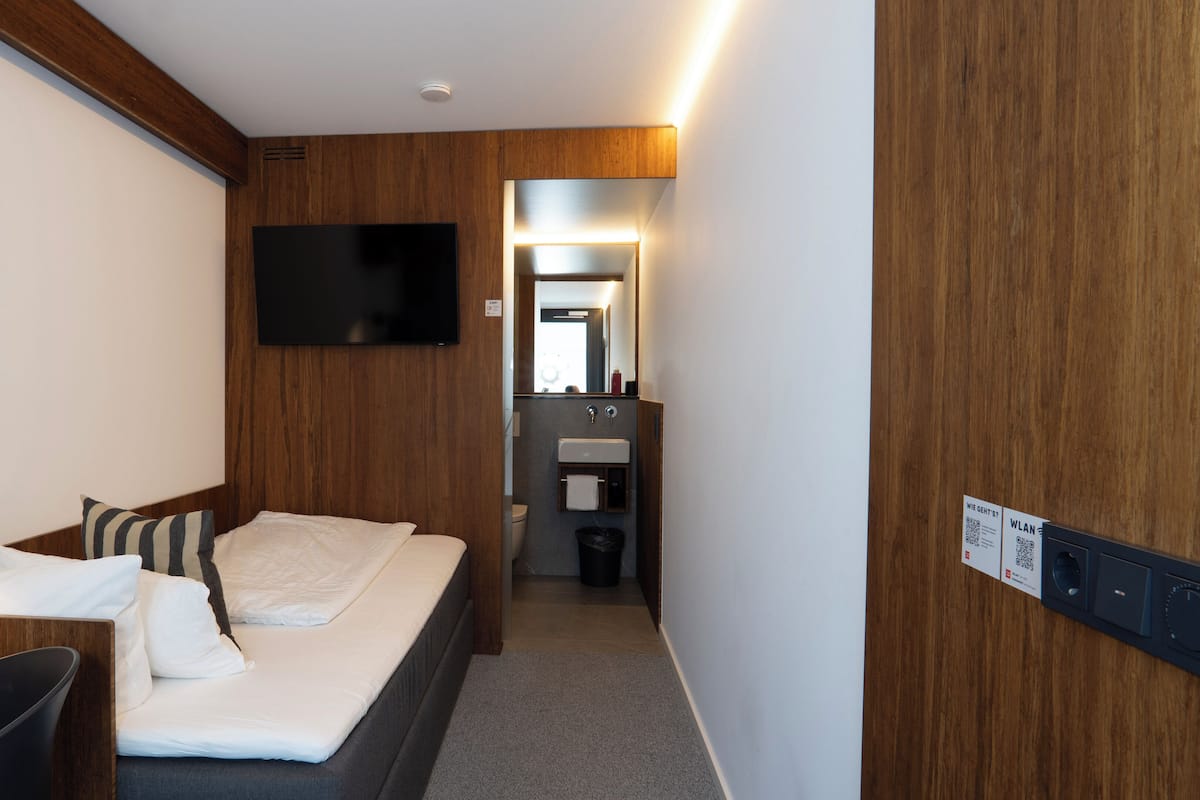 A hotel room with a bed, a desk, a chair, a TV, a mirror, a sink, and a trash bin.