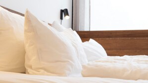 Una cama bien hecha con sábanas y almohadas blancas, un cabecero de madera y una lámpara fijada a la pared.
