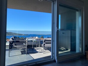 Un balcón con vistas al mar y a las colinas, con puerta de cristal, mobiliario exterior y suelo de baldosas.