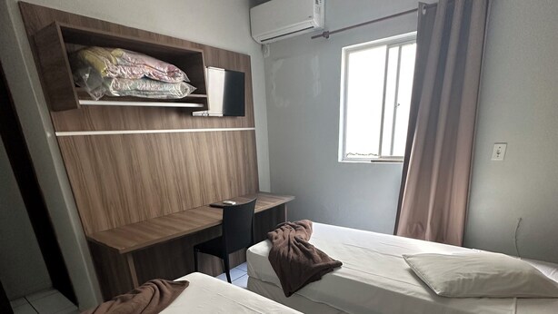 A hotel room with two single beds, a desk with a chair, a TV, and an air conditioning unit. - Renata Palace Hotel (Tubarao)
