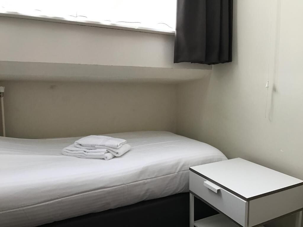 Ein kleines Zimmer mit einem Einzelbett, einem weißen Nachttisch und einem Fenster mit schwarzem Vorhang.