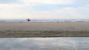 A beach with a person holding a surfboard, calm water, and a clear sky. - Hotel Carillon (Haarlem)