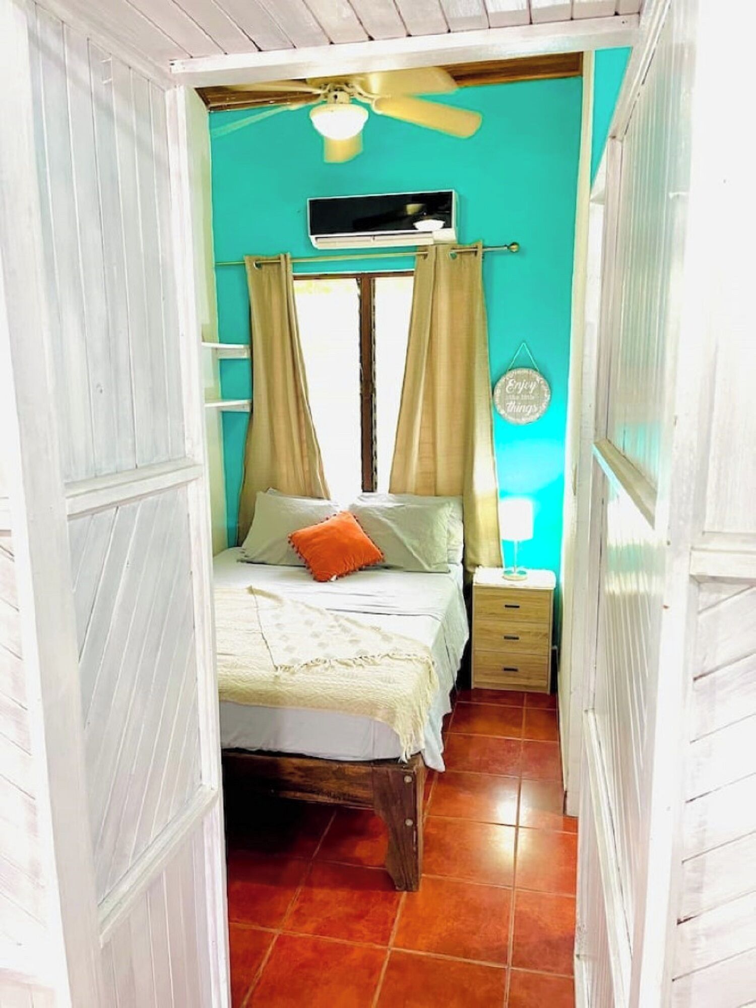 A narrow room with a bed, a nightstand, a ceiling fan, and a window with curtains.