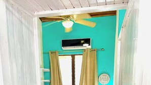 Ein schmaler Raum mit einem Bett, einem Nachttisch, einem Deckenventilator und einem Fenster mit Vorhängen.
