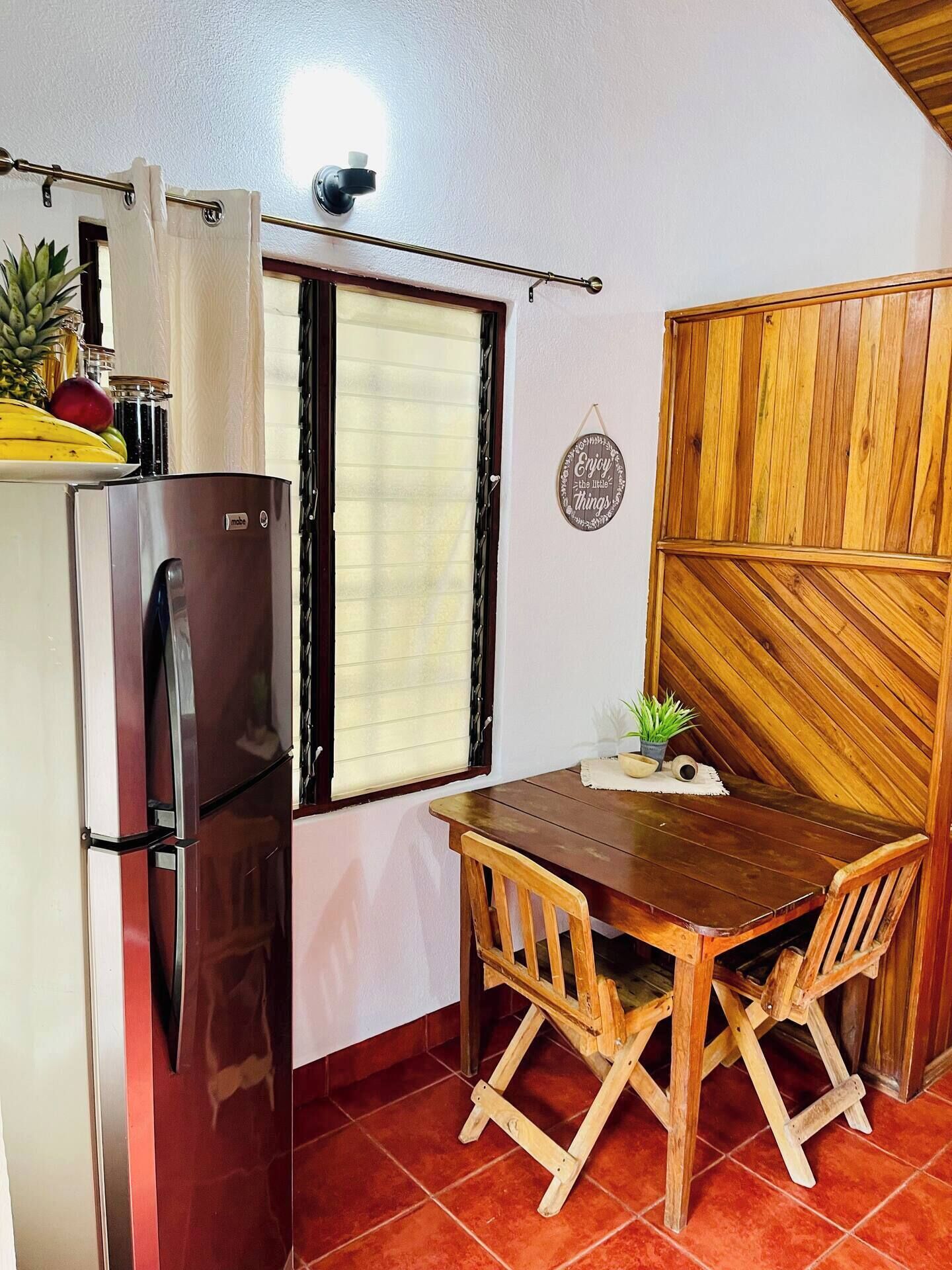 A compact kitchen with a refrigerator, a window with blinds, a wooden dining table with chairs, and a wall-mounted sign.