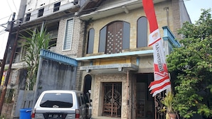 A two-story building with a traditional architectural style, featuring a prominent entrance with decorative ironwork and a large wooden door. A flag is displayed prominently on the facade, and a silver van is parked in front of the building. - OYO Life  92842 Homestay Balearjosari (Malang)