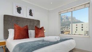 A modern bedroom with a large bed, two red pillows, a grey headboard, and a view of a mountain through a large window. - Luxury On Loop with Table Mountain Views
 (Cape Town)