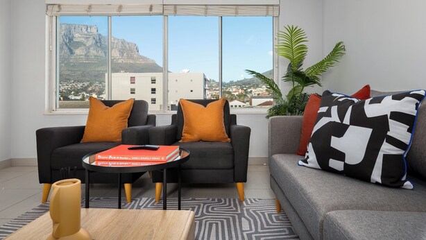 A modern living room with a large window, two sofas with orange and black cushions, a small table with a book, and a potted plant. - Luxury On Loop with Table Mountain Views
 (Cape Town)