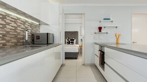 A modern kitchen with a built-in oven, a microwave, and a coffee machine. There is a bed visible through an open door in the background. - Luxury On Loop with Table Mountain Views
 (Cape Town)
