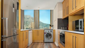 A modern kitchen with wooden cabinets, a washing machine, and a view of a cityscape through large windows. - Modern & Open 2BR with Table Mountain & Lions Head Views
 (Cape Town)