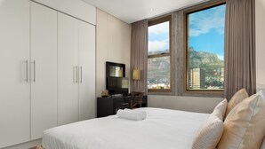 A hotel room with a large bed, a TV, a desk, and a view of a cityscape through a window. - Modern & Open 2BR with Table Mountain & Lions Head Views
 (Cape Town)