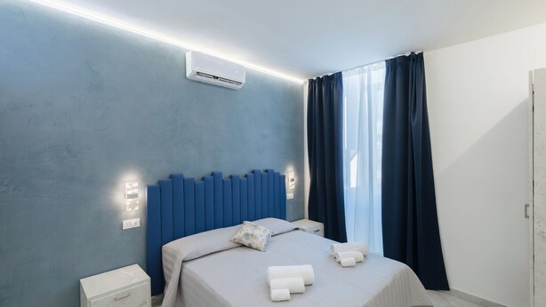 Une chambre moderne avec un mur d’accent bleu, une table de chevet blanche, un lit bien fait avec des serviettes, et une fenêtre avec des rideaux.