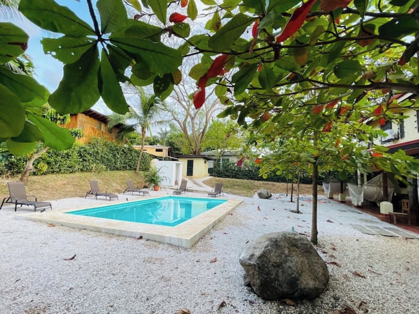 A swimming pool surrounded by a gravel area with lounge chairs and a large rock.