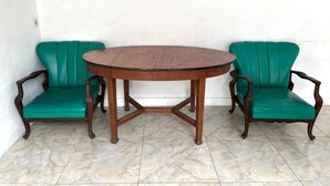 Une table ronde en bois, dotée d’une structure en croisillons, est entourée de deux chaises vertes à revêtement rembourré et aux accoudoirs en bois.