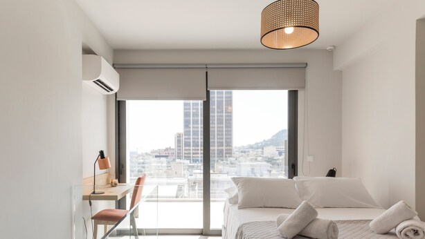 A modern hotel room with a city view, a desk with a lamp, a bed with pillows, and a hanging light fixture. - Cubes Living Athens, Duplex Apartment 801
 (Athina)