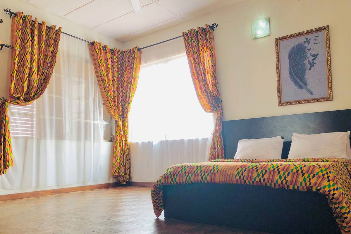 Una habitación de hotel con cama, cortinas, cabecera y un cuadro colgado en la pared.