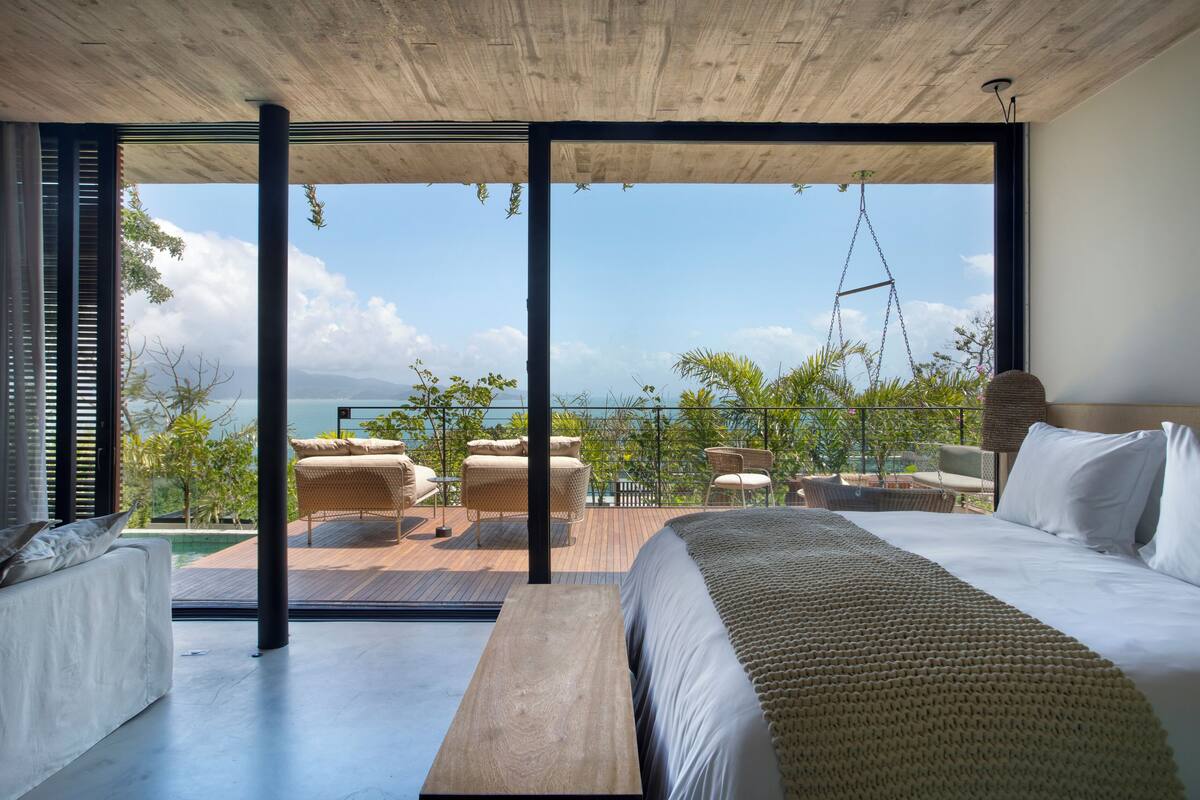 A bedroom with a large bed, a wooden bench, and a view of the ocean through floor-to-ceiling windows.