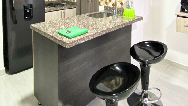 A compact kitchen with a refrigerator, microwave, and a sink. There are two bar stools and a green cutting board on the counter. - Apartahotel222 Apto 401 (Pereira)