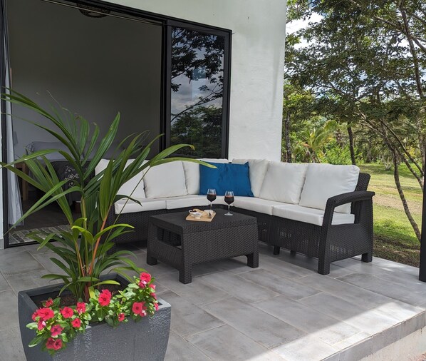 Un área de estar moderna al aire libre con un sofá blanco y una mesa de centro de mimbre negro, rodeada de plantas en macetas y vistas del exterior.
