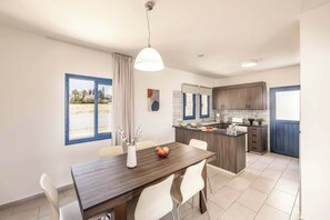 A modern kitchen with a central island, white chairs around a dining table, and a view of the outdoors through a window. - Coral Olympus Apollo (Peyia)