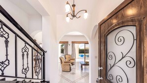 A grand staircase with ornate railings, a chandelier, and a view into a living area with a pool. - Villa Kyveli 2 - Three Bedroom Villa, Sleeps 6 (Peyia)