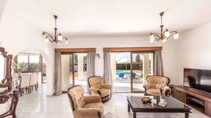 A spacious living room with a large TV, two patterned armchairs, a dining area with a table and chairs, and a view of a swimming pool through the glass doors. - Villa Kyveli 2 - Three Bedroom Villa, Sleeps 6 (Peyia)
