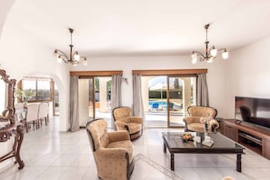 Ein geräumiges Wohnzimmer mit großem Fernseher, zwei gemusterten Sesseln, einer Essecke mit Tisch und Stühlen sowie Blick auf einen Swimmingpool durch Glastüren.