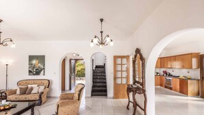 A spacious living room with a sofa, a coffee table, and a chandelier. There is a staircase leading to a kitchen area with wooden cabinets and a tiled floor. - Villa Kyveli 2 - Three Bedroom Villa, Sleeps 6 (Peyia)