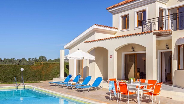 A pool area with lounge chairs and a table, a building with a tiled roof, and a clear sky. - Villa Kyveli 2 - Three Bedroom Villa, Sleeps 6 (Peyia)