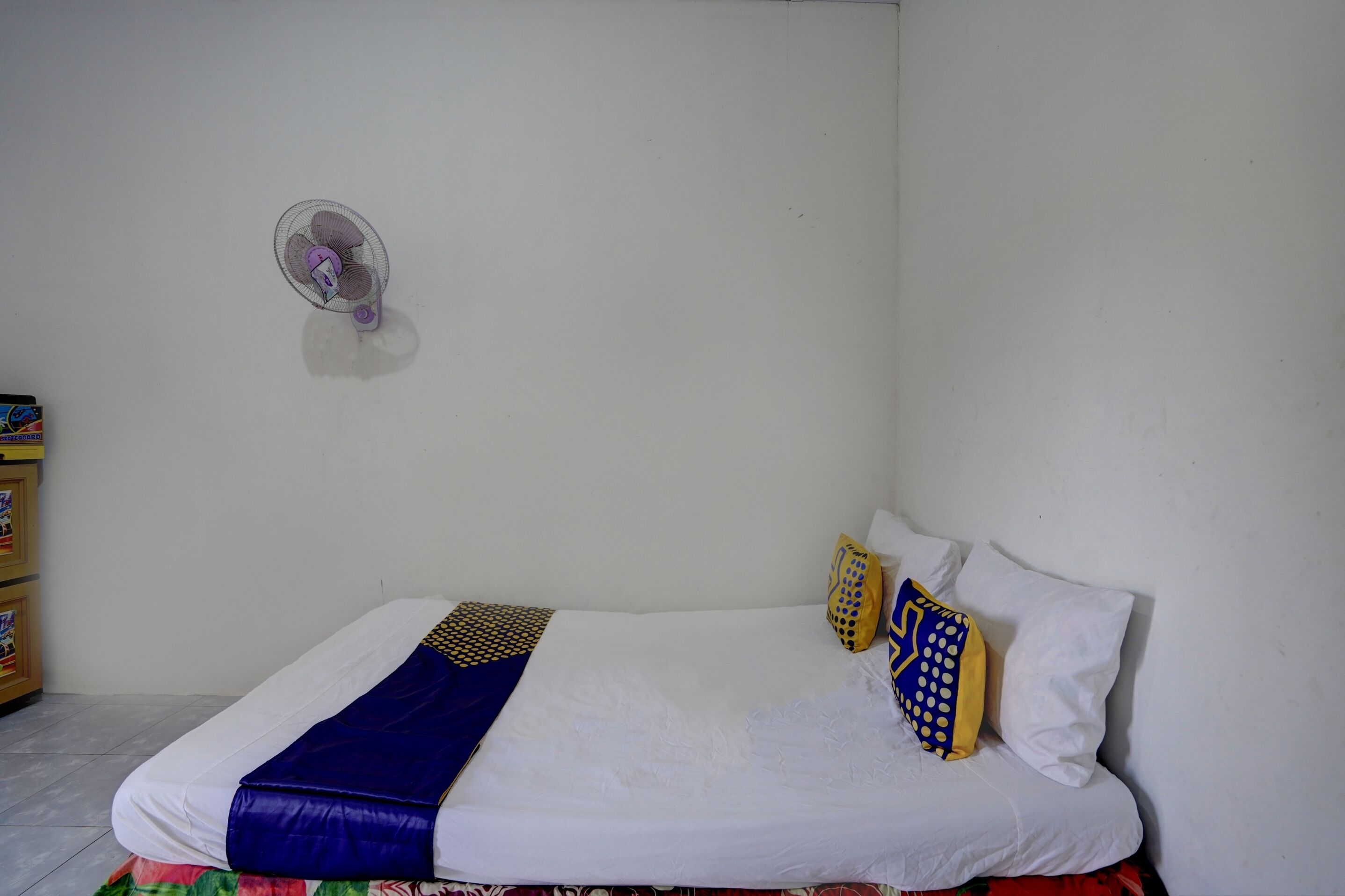 A single bed with white bedding, a blue and yellow blanket, and patterned pillows. A small wall fan is mounted on the wall.