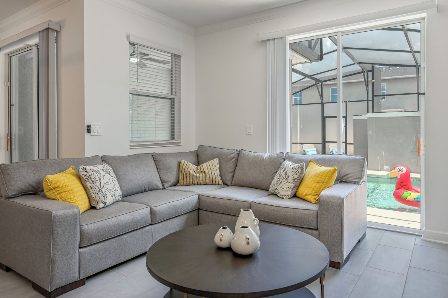 A modern living room with a grey sectional sofa, a round coffee table, and a view of a pool through large glass doors.