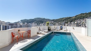 A rooftop pool with white lounge chairs and a table, surrounded by buildings and a hill in the background.