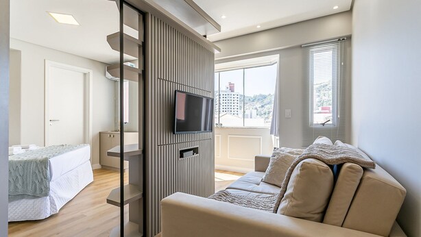 A modern living room with a sofa, a TV mounted on a wall, and a balcony with a view of the city.