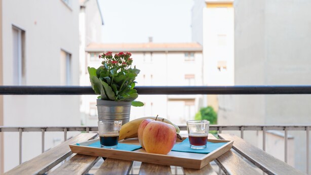 A wooden table with a tray holding a peach, a banana, and two glasses of dark liquid, possibly coffee or tea, on a balcony with a metal railing and a potted plant. - Milan Hub - Sempione, Citylife, MiCo (Milano)
