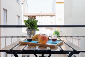A wooden table with a tray holding a peach, a banana, and two glasses of dark liquid, possibly coffee or tea, on a balcony with a metal railing and a potted plant.