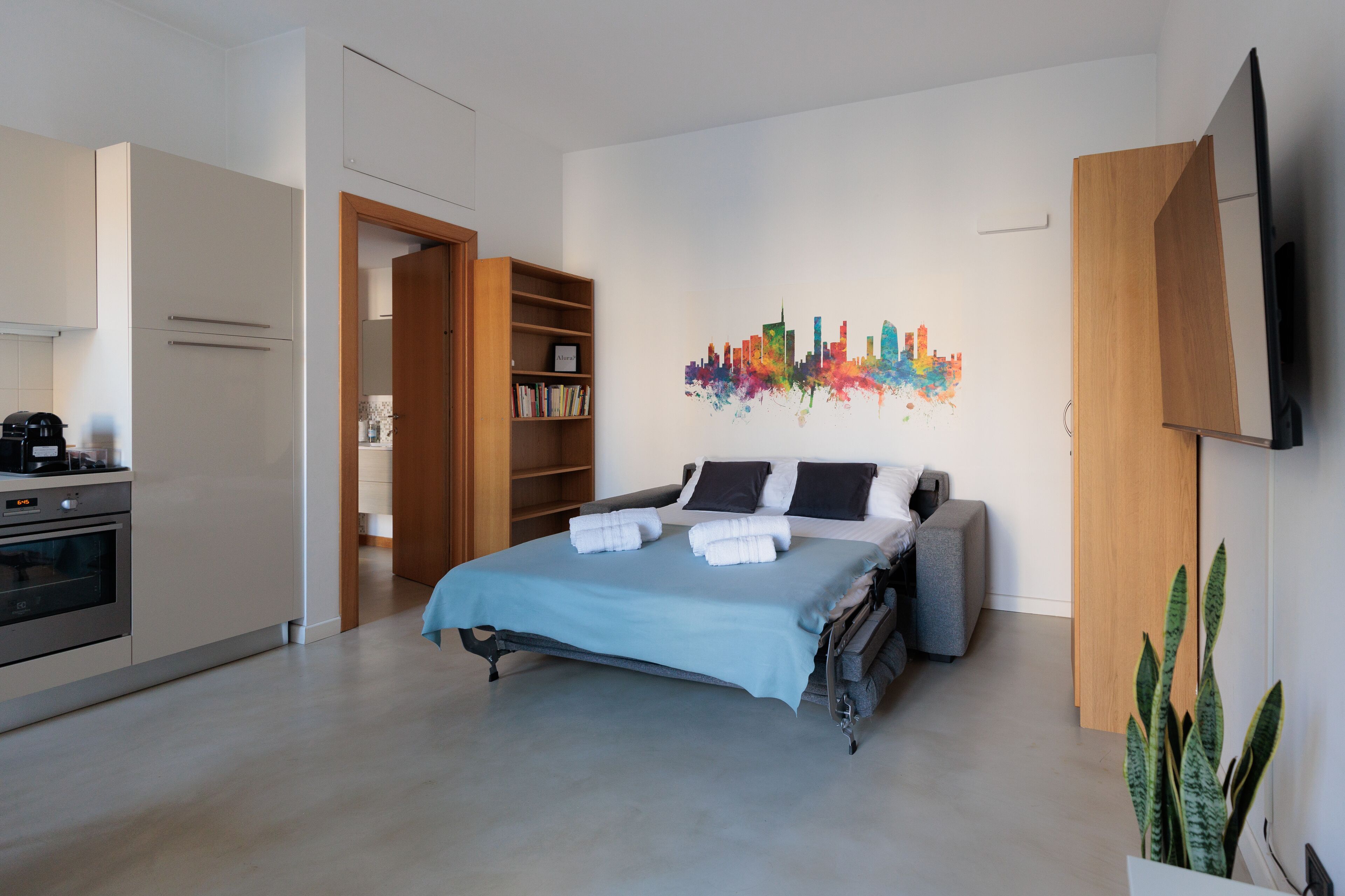 A modern hotel room with a bed, a city skyline artwork on the wall, a television, and a kitchen area.