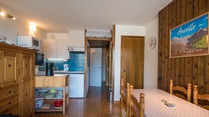 Una habitación compacta con cama, cabecero de madera, una pequeña kitchenette con microondas, hervidor y refrigerador, y un comedor con mesa y sillas.