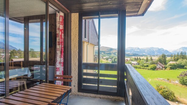 Un balcón con mobiliario de madera, vista a un paisaje montañoso y cielo despejado.