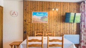Un comedor con una mesa y sillas de madera, un reloj en la pared, un cuadro enmarcado de un paisaje montañoso y un televisor colgado en la pared.