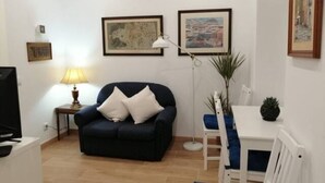 Una sala de estar con un sofá azul oscuro, una lámpara blanca, una mesita y una silla. Hay dos cuadros en la pared.