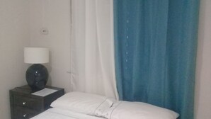 A hotel room with a bed, white bedding, a nightstand with a lamp, and a blue curtain. - Tranquille2, Jamaica vacation Kingston/St. Andrew
 (Kingston)