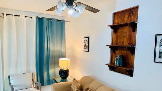 A living room with a beige sofa, patterned cushions, a wicker chair, a wooden shelf, and a ceiling fan. - Tranquille2, Jamaica vacation Kingston/St. Andrew
 (Kingston)