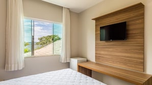 Quarto com uma janela grande, uma cama, uma TV fixada em um painel de madeira e um armário pequeno branco.