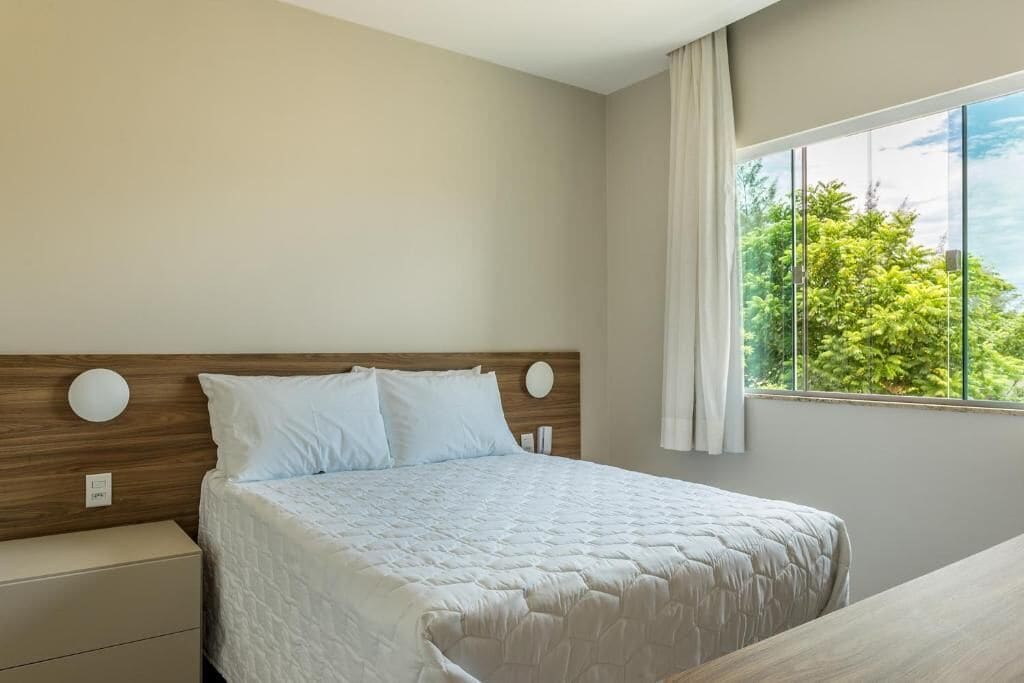 A bedroom with a bed, a nightstand, a window with curtains, and a view of trees.