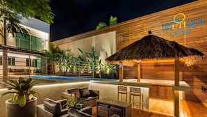 Uma área de piscina moderna ao ar livre, com mobiliário de lounge, um bar e um guarda-sol de palha.