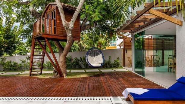 Uma casa na árvore de madeira presa a uma árvore, uma cadeira suspensa, uma piscina e uma área de estar moderna ao ar livre.