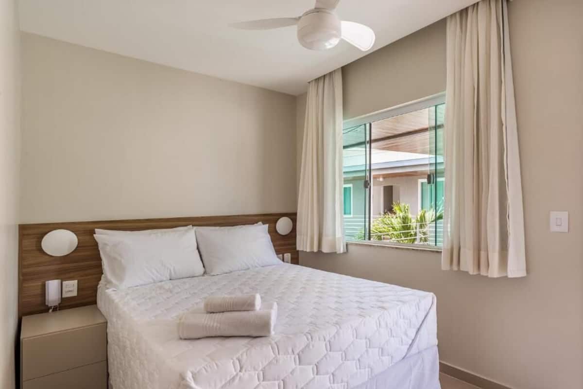 A neatly made bed with a quilted cover and two pillows, a bedside table with a lamp, a ceiling fan, and a window with curtains.