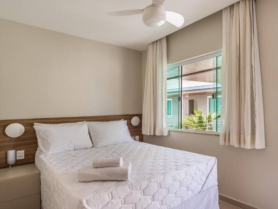 A neatly made bed with a quilted cover and two pillows, a bedside table with a lamp, a ceiling fan, and a window with curtains.