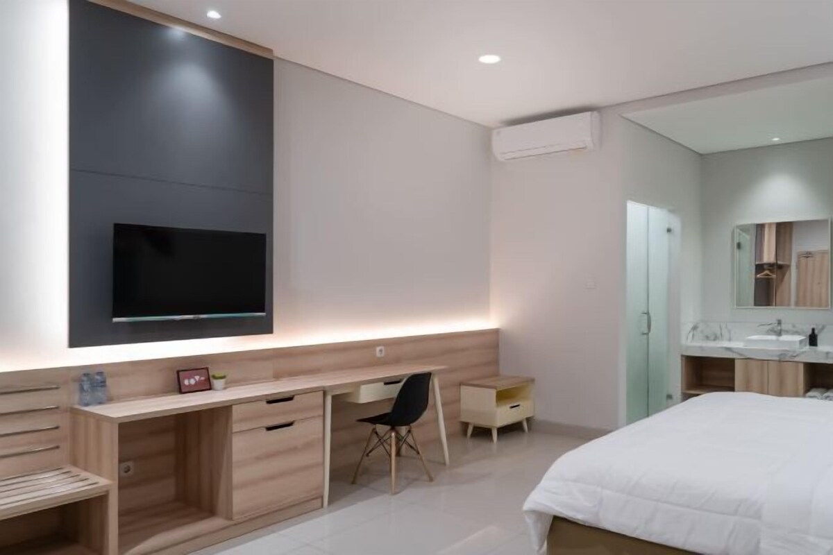 A modern bedroom with a bed, a desk, a chair, a TV, and a bathroom with a mirror and sink.
