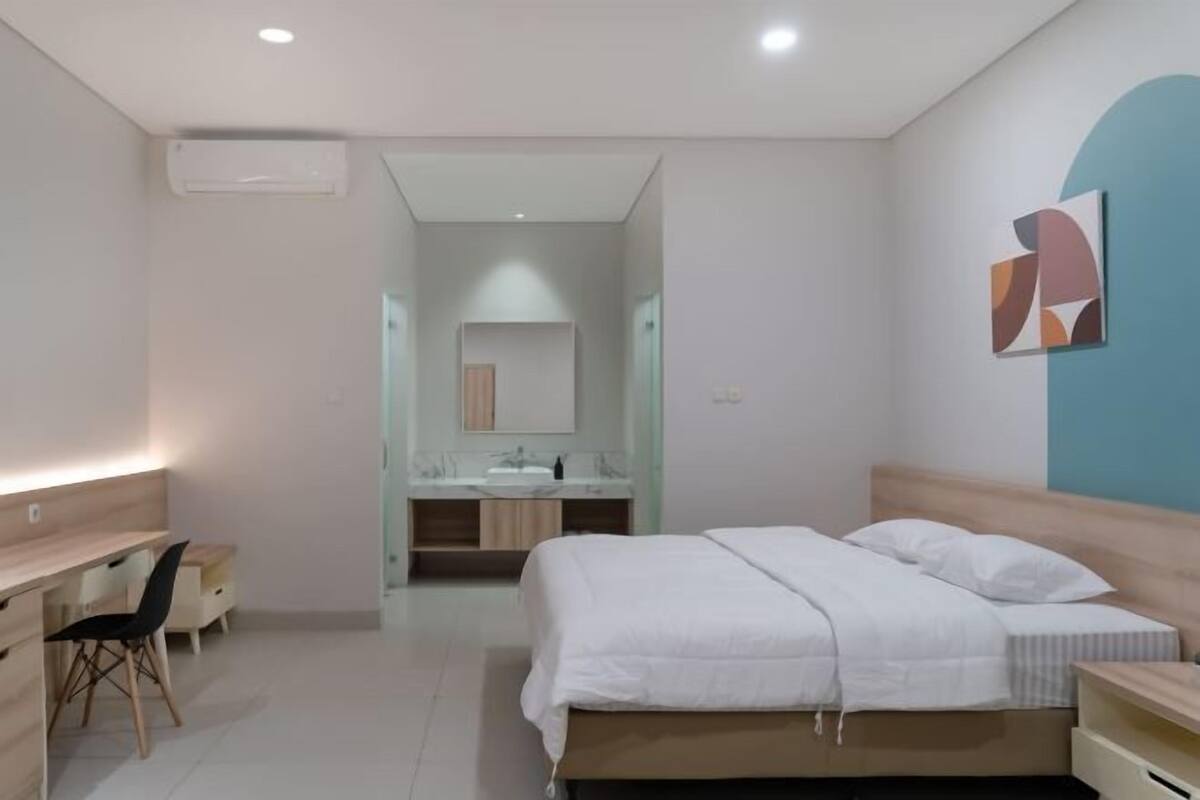 A modern bedroom with a bed, desk, chair, and a bathroom visible in the background.